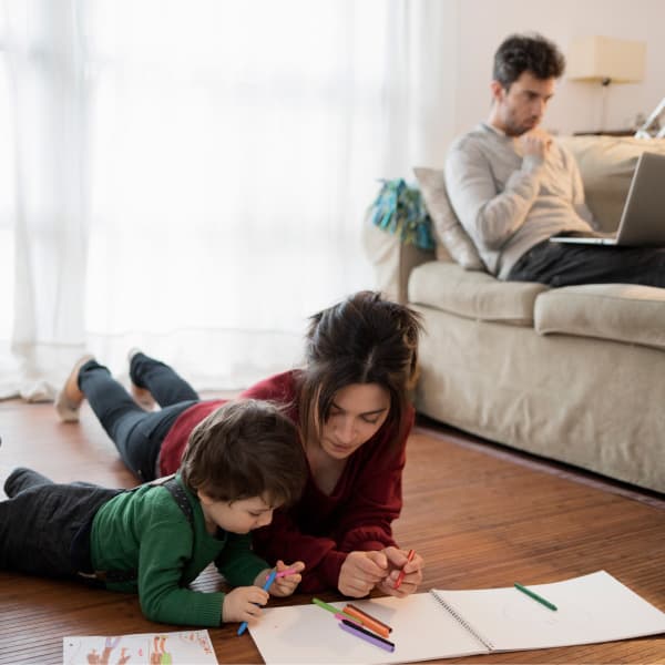 Woman and young child coloring on the floor with men on laptop on the couch in the background.
