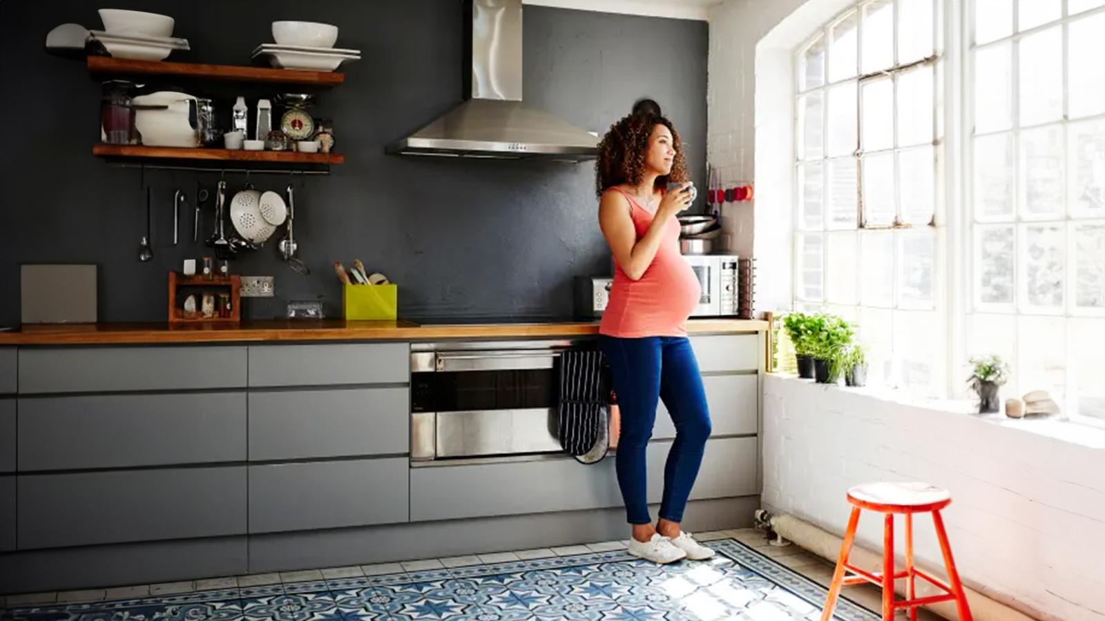 Pregnant woman drinking coffee and looking out of kitchen window.